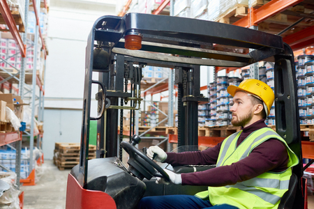 Side view portrait of young warehouse worker sitting inside forklift moving goods from tall storage shelves, copy spaceの写真素材