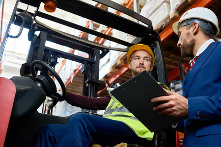 Low angle portrait of mature businessman talking to warehouse worker sitting in forklift car, copy spaceの写真素材