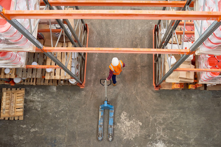 Top view background image of tall shelf rows in modern warehouse with worker wearing hardhat pulling empty cart in aisle, copy spaceの写真素材