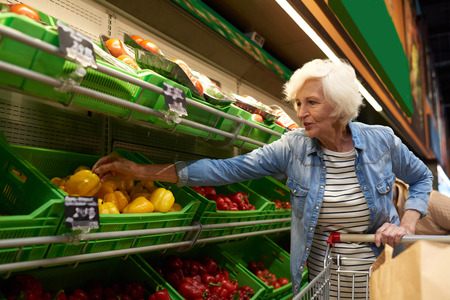 Portrait of modern senior woman with shopping cart  choosing fruits and vegetables in supermarket while enjoying grocery shopping, copy spaceの写真素材
