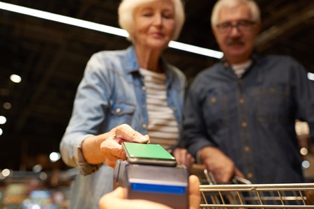 Waist up portrait of modern senior couple buying groceries in supermarket paying with NFC payment via smartphone, focus on hand, copy spaceの写真素材