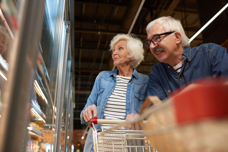 Low angle portrait of modern senior couple grocery shopping in supermarket, smiling happily while choosing frozen foods standing by freezers , copy spaceの写真素材