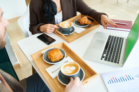 High angle crop shot of two business people, man and woman, enjoying coffee break while working in cafe, focus on coffee cups and croissants on wooden trays by laptop with green screenの写真素材