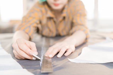 Close up of unrecognizable female tailor tracing fabric with white chalk while sewing custom made...
