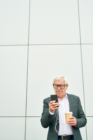 Aged businessman with cup using smartphone near buildingの写真素材