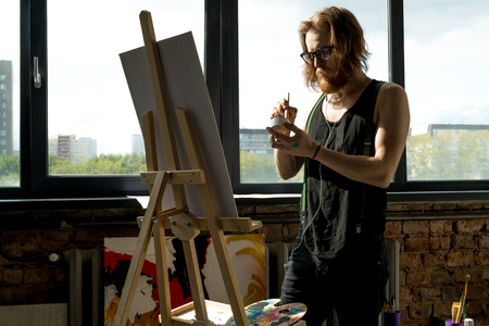 Portrait of contemporary male artist painting pictures standing by easel in art studio lit by sunlight, copy spaceの写真素材