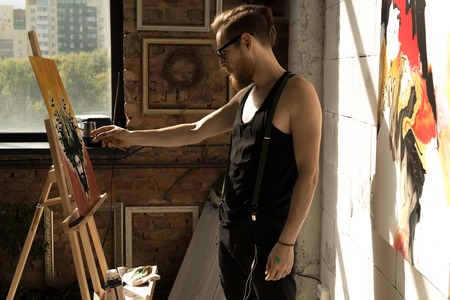 Side view portrait of handsome male artist painting picture on easel in art studio lit by sunlight, copy spaceの写真素材