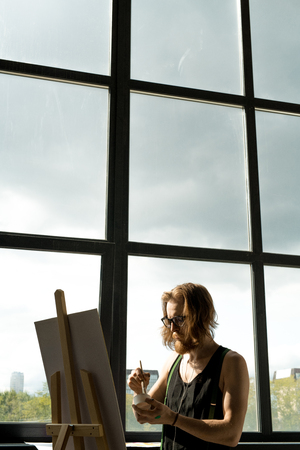 Waist up portrait of contemporary male artist painting pictures standing by easel in art studio lit by sunlight, copy spaceの写真素材