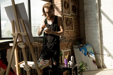 Portrait of contemporary long haired artist painting pictures standing by easel in art studio lit by sunlight, copy spaceの写真素材
