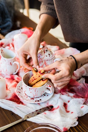 Close-up of unrecognizable girl standing at table with tea crockery and gauze and adjusting witch finger cookie in cup while making Halloween decor  in workshopの写真素材
