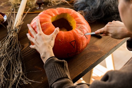 Close-up of unrecognizable woman sitting at table and cutting eye on pumpkin while making jack-o-lantern in workshopの写真素材