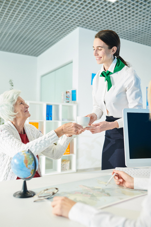 Secretary giving cup of tea to senior lady in travel agency offiの写真素材