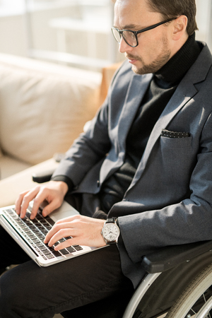 Serious young businessman in eyeglasses sitting in wheelchair and typing on his laptop which is on his kneesの写真素材
