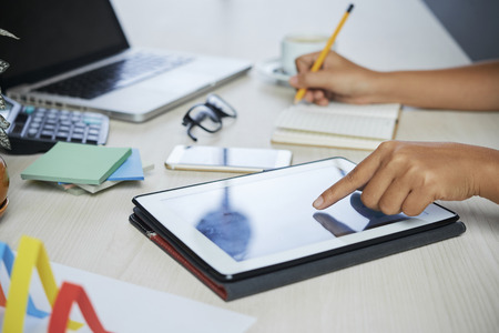 Office worker reading financial report on tablet computer and writing in planner