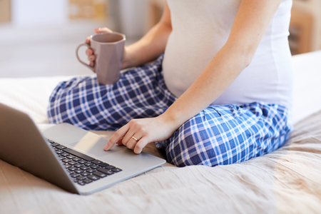 Closeup of pregnant woman using laptop siting on bed in warm sunlight, copy spaceの写真素材
