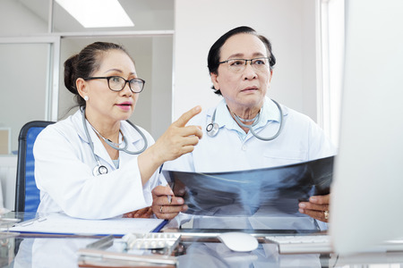 Serious concerned Vietnamese doctors holding lungs x-ray and reading results of its interpretation on computer screenの写真素材