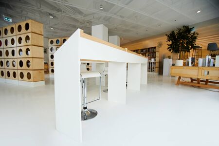 White angled tables and modern high chairs placed in center of bookstore room, creative bookcases in...