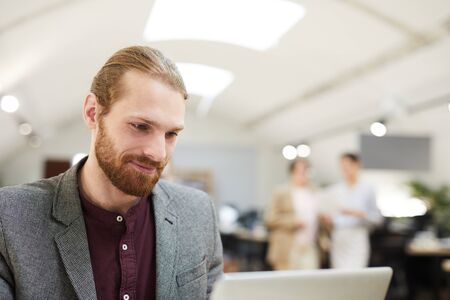 Portrait of handsome bearded businessman smiling while looking at laptop screen, copy space