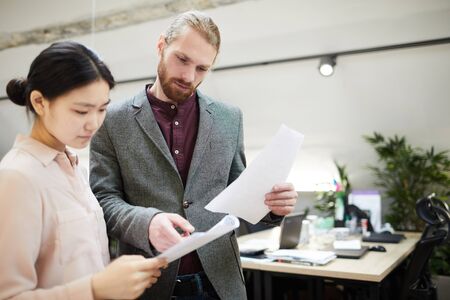 Portrait of handsome businessman discussing documents with Asian colleague while standing in office, copy spaceの写真素材