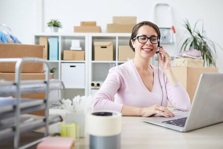 Smiling young helpdesk operator in headset talking in microphone while sitting in front of laptop by deskの写真素材