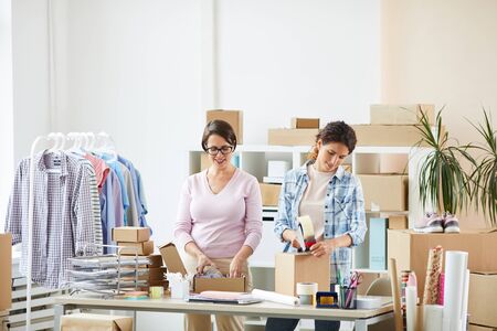 Young female colleagues standing by desk while making parcels for their online clients and sealing...