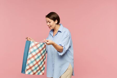Young excited female in casualwear looking at surprise in open paperbag while posing in isolation