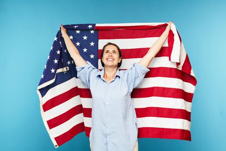 Happy young casual woman raising hand while holding large American flag on blue backgroundの写真素材
