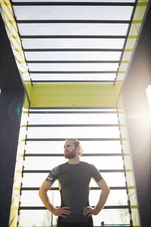 Serious young motivated sportsman with red beard standing at modern sports equipment outdoors