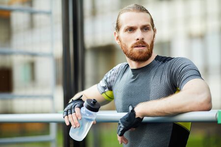 Pensive young bearded sportsman in sweaty sports shirt leaning on bar and drinking water while...