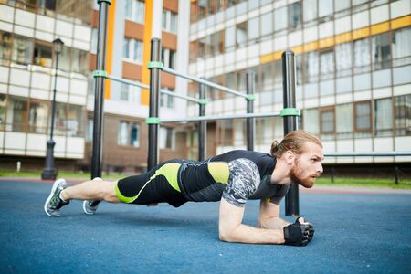 Serious motivated young man with beard and ponytail leaning on elbows while doing plank outdoors
