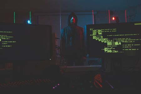 Serious bearded cyber-terrorist in hoodie standing in computer room with alarm system, computer monitors with codes on foregroundの写真素材
