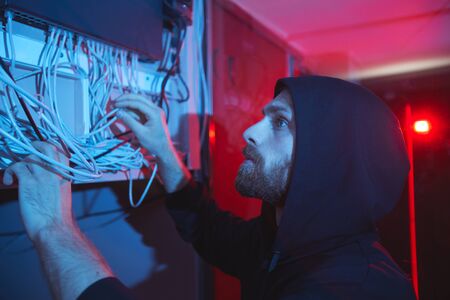 Serious concentrated young bearded network specialist in hoodie standing in dark room and examining...