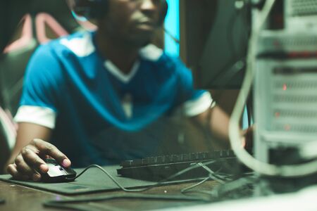 Close-up of black man in tshirt sitting at table with cables and clicking with mouse while shooting in computer gameの写真素材