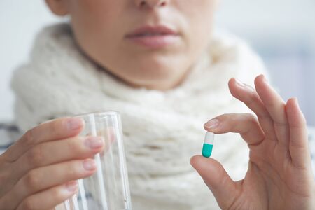 Close-up of young woman in warm scarf around neck holding glass of water and taking antiviralsの写真素材