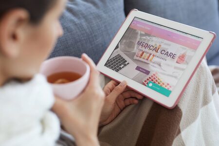 Close-up of sick woman sitting under blanket on sofa and using tablet while reading how to cure coldの写真素材