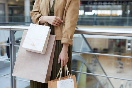 Close-up of stylish lady in jacket and dress standing at railing in mall and holding shopping bagsの写真素材
