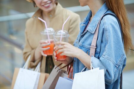 Close-up of positive ladies with shopping bags drinking sweet cocktails and chatting in mallの写真素材
