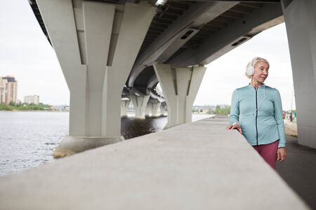 Retired active female in sportswear and headphones listening to music while having rest under large bridge at leisureの写真素材