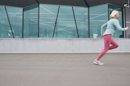 Contemporary active mature sportswoman running down concerete road along building of modern design...