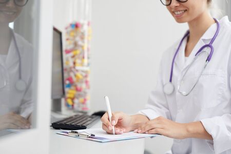 Mid-section portrait of smiling female doctor writing on clipboard while sitting at desk in office, copy spaceの写真素材