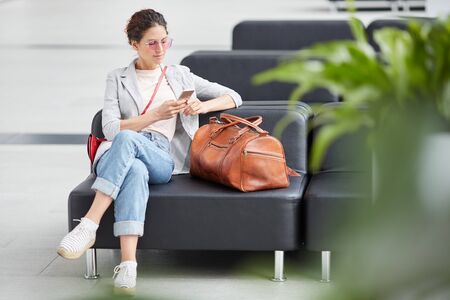 Pensive young girl in pink sunglasses sitting on leather sofa in airport and using gadget while waiting for departureの写真素材