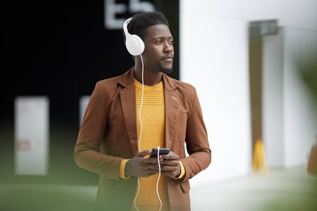 Pensive dreamy young black man in casual jacket standing in lobby and listening to audiobook in...
