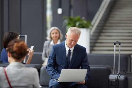 Content handsome gray-haired businessman in formal suit sitting on sofa and examining data on laptop while waiting for boardingの写真素材