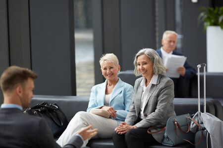 Cheerful mature business colleagues in jackets sitting on sofa in waiting area of airport and chatting while waiting for boardingの写真素材