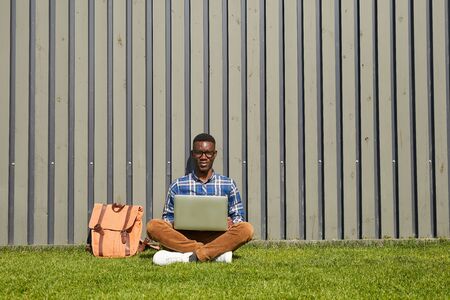Full length portrait of African IT student looking at camera while using laptop outdoors in college...