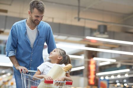 Portrait of happy young father grocery shopping in supermarket and smiling at little girl sitting in shopping cart, copy spaceの写真素材
