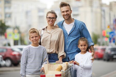 Portrait of contemporary family with two kids posing with shopping cart at parking lot outdoors, copy spaceの写真素材