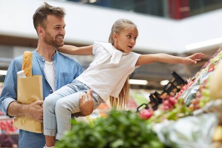 Portrait of cute little girl reaching for vegetables while enjoying grocery shopping with dad at farmers market, copy spaceの写真素材