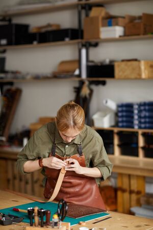 Waist up portrait of woman artisan making leather belt in shop lit by sunlight, copy spaceの写真素材