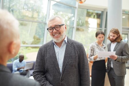 Portrait of smiling senior businessman wearing glasses talking to colleague standing in modern...